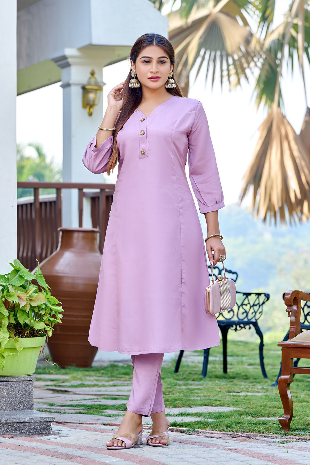 Woman in a light purple outfit standing outdoors with plants and furniture in the background